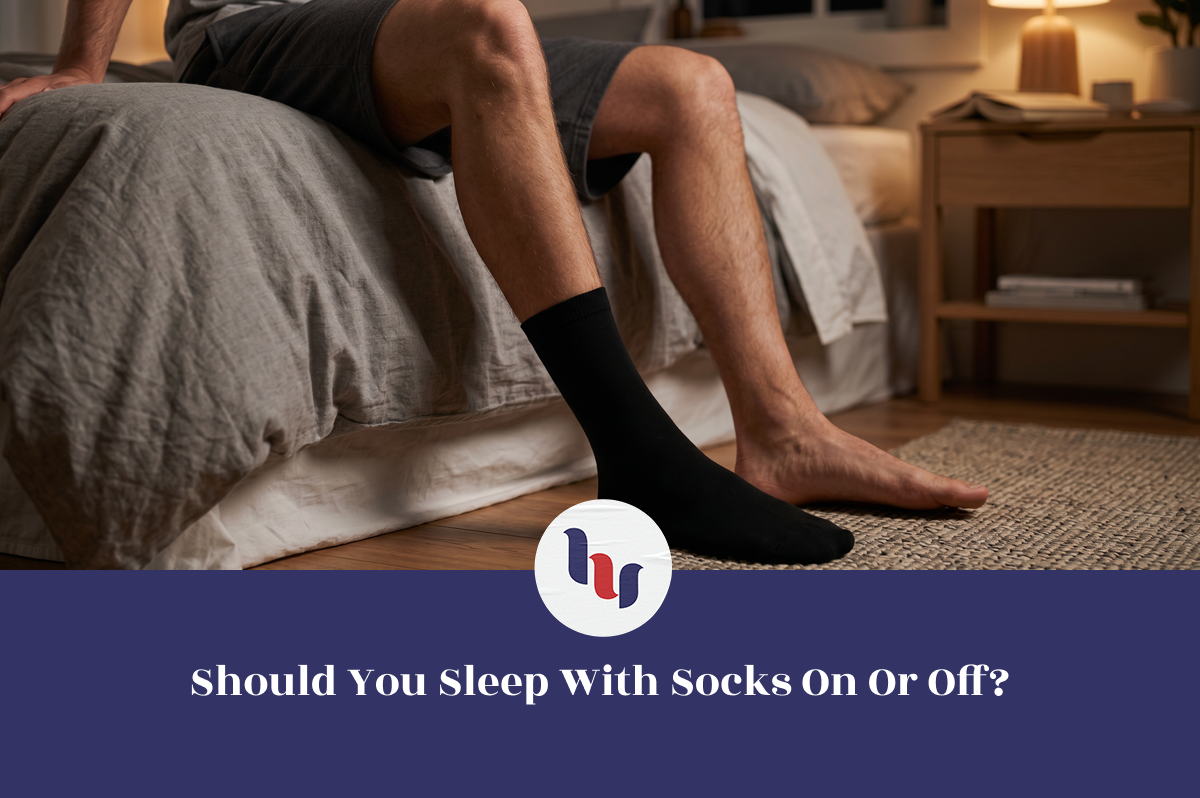 Person sitting on the edge of a bed at night with one foot wearing a black sock and the other foot bare, illustrating the question of whether sleeping with socks on or off is better for comfort and sleep quality.