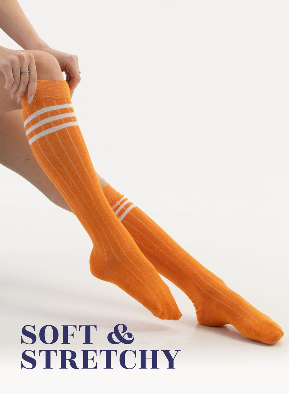 Orange knee-high socks with white stripes worn by a person on a light background, with text 'SOFT & STRETCHY'.