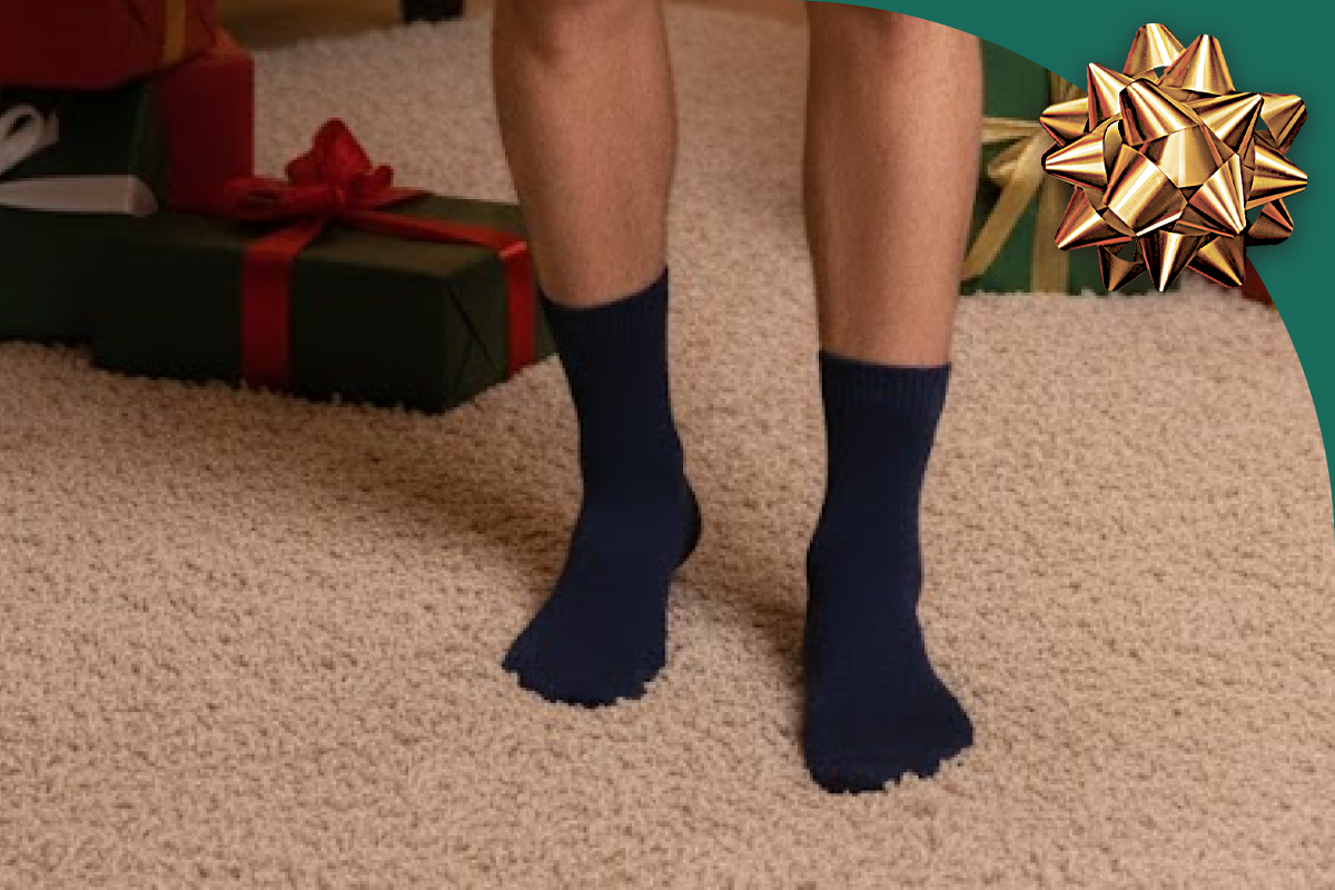 A man standing on a carpet wearing navy Hugh Ugoli socks with Christmas gift boxes in the background.
