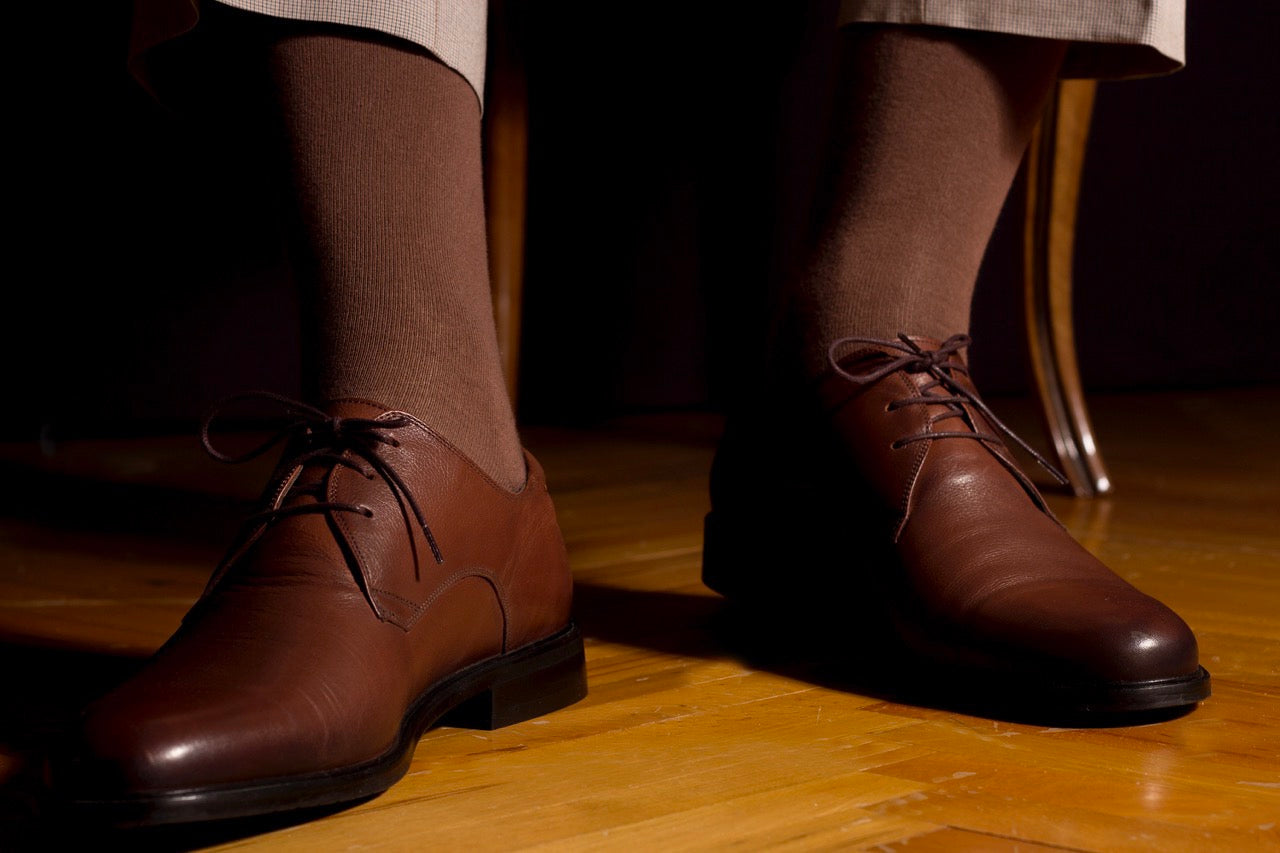 A person wearing light brown dress shoes paired with dark brown socks, seated with their feet flat on a wooden floor, likely in an indoor setting with warm lighting.
